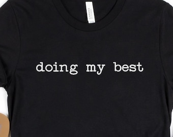 May include: Black t-shirt with the text "doing my best" in white lettering. The shirt is shown with a pair of brown flip flops and denim shorts.