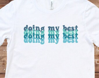 May include: White t-shirt with the text "doing my best" repeated four times in a gradient of blue and green colors.