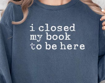 May include: Denim blue sweatshirt with the text "i closed my book to be here" in white. The long-sleeved top is a casual style, suitable for everyday wear. The sweatshirt is a solid color.