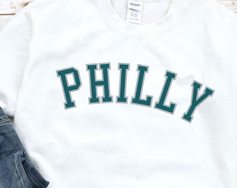 May include: White sweatshirt with a teal blue and gray arched graphic that says "PHILLY". The sweatshirt is paired with blue jeans and white sneakers.