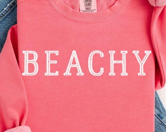 May include: A watermelon-colored sweatshirt with the word "BEACHY" in white block letters. The sweatshirt is folded over a pair of blue denim jeans. The sleeves are cuffed, and the fabric appears soft and comfortable.