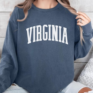 May include: Denim blue sweatshirt with the word "VIRGINIA" in white, arched lettering. The sweatshirt has long sleeves and a crew neck. The image shows the sweatshirt on a person.