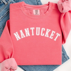 May include: A pink sweatshirt with the word "NANTUCKET" in white text on the front. The sweatshirt is folded and laid on top of a pair of blue jeans.