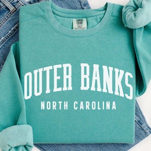 May include: A seafoam green sweatshirt with the words "OUTER BANKS NORTH CAROLINA" in white block letters. The sweatshirt is folded over a pair of blue jeans.