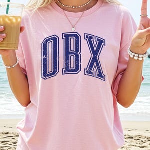 May include: A light pink t-shirt with the letters "OBX" in a distressed navy blue font. The shirt has a crew neck and short sleeves. The background shows a beach and ocean.