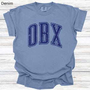 May include: A light blue t-shirt with the word "OBX" in navy blue, distressed lettering. The shirt has short sleeves and a crew neck. The words "Washed Denim" are in the top left corner.