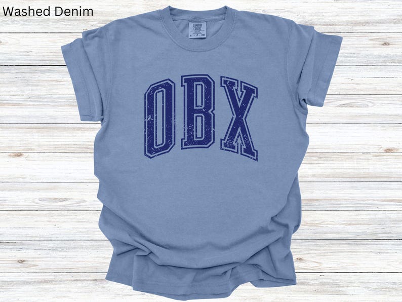 May include: A light blue t-shirt with the word "OBX" in navy blue, distressed lettering. The shirt has short sleeves and a crew neck. The words "Washed Denim" are in the top left corner.