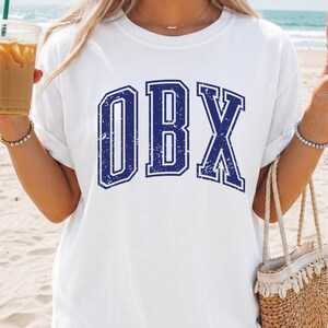 May include: White t-shirt with the blue letters "OBX" in a collegiate font. The shirt is worn on a beach with a straw bag and a drink in hand.