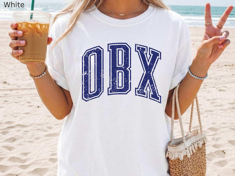 May include: White t-shirt with the blue letters "OBX" in a collegiate font. The shirt is worn on a beach with a straw bag and a drink in hand.