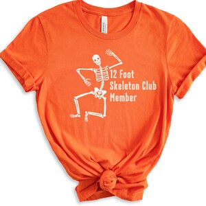 May include: Orange t-shirt with a white skeleton graphic and the text "12 Foot Skeleton Club Member".