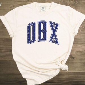 May include: Ivory-colored t-shirt with the letters "OBX" in a distressed navy blue font. The shirt is knotted at the bottom and has rolled-up sleeves. The text "ivory" is in the top left corner.