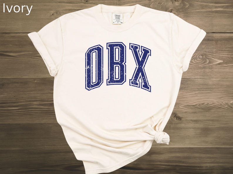 May include: Ivory-colored t-shirt with the letters "OBX" in a distressed navy blue font. The shirt is knotted at the bottom and has rolled-up sleeves. The text "ivory" is in the top left corner.