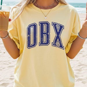 May include: Butter yellow t-shirt with the text "OBX" in distressed navy blue lettering. The shirt is a relaxed fit and is being worn on a beach. A clear plastic cup with iced coffee is held in the left hand.