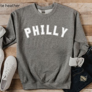 May include: A gray heather sweatshirt with the word "PHILLY" printed in white on the front. The sweatshirt is laid flat on a wooden surface.