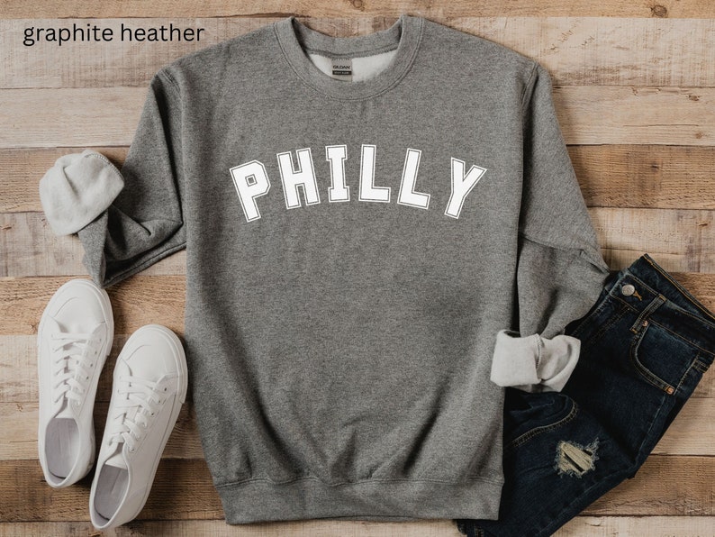 May include: A gray heather sweatshirt with the word "PHILLY" printed in white on the front. The sweatshirt is laid flat on a wooden surface.
