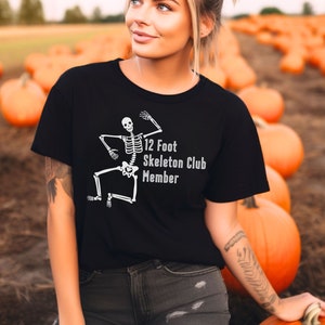 May include: A black t-shirt with a white skeleton graphic and the text "12 Foot Skeleton Club Member". The person wearing the shirt is standing in a field of pumpkins.