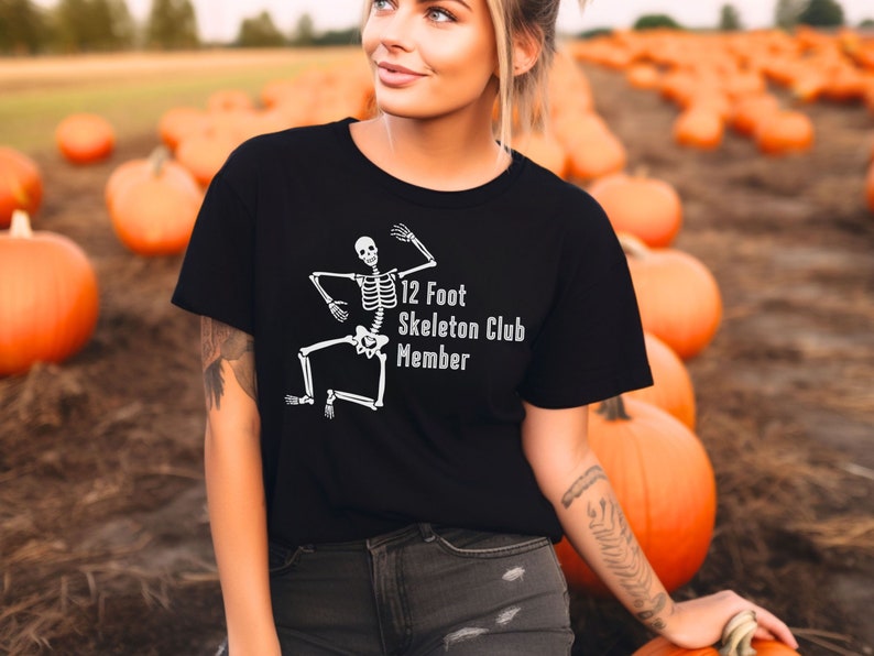 May include: A black t-shirt with a white skeleton graphic and the text "12 Foot Skeleton Club Member". The person wearing the shirt is standing in a field of pumpkins.
