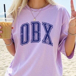 May include: A light purple t-shirt with the letters "OBX" in navy blue, with a distressed, vintage look. The shirt is worn on a beach with a drink in hand. The word "Orchid" is in the top left corner.
