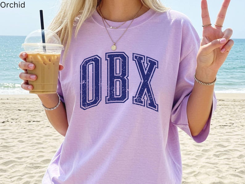 May include: A light purple t-shirt with the letters "OBX" in navy blue, with a distressed, vintage look. The shirt is worn on a beach with a drink in hand. The word "Orchid" is in the top left corner.