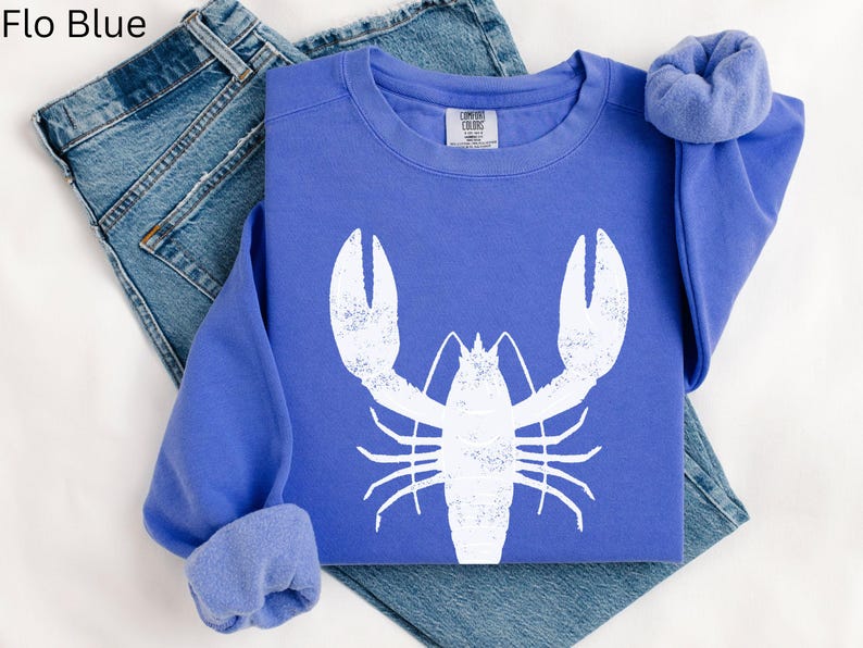 May include: A blue sweatshirt with a white lobster graphic. The sweatshirt is folded and laid on a white surface with a pair of blue jeans.