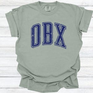 May include: A light green t-shirt with the letters "OBX" in distressed navy blue, arched across the front. The shirt has a crew neck and short sleeves. The word "Bay" is in the top left corner.