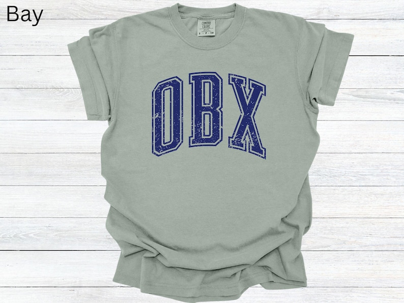 May include: A light green t-shirt with the letters "OBX" in distressed navy blue, arched across the front. The shirt has a crew neck and short sleeves. The word "Bay" is in the top left corner.