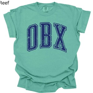 May include: A mint green t-shirt with the word "OBX" in navy blue, distressed collegiate-style lettering. The shirt is folded, showing the sleeves and the front of the garment. The text "Island Reef" is in the top left corner.