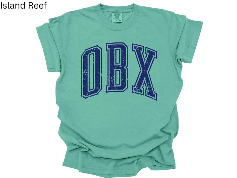 May include: A mint green t-shirt with the word "OBX" in navy blue, distressed collegiate-style lettering. The shirt is folded, showing the sleeves and the front of the garment. The text "Island Reef" is in the top left corner.