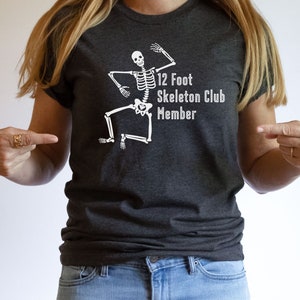 May include: A dark gray t-shirt with a white skeleton graphic and the text "12 Foot Skeleton Club Member".
