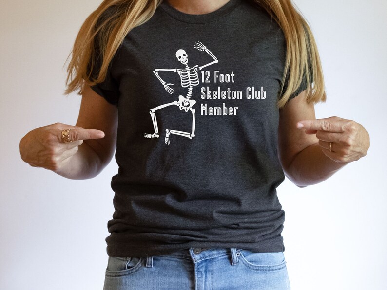 May include: A dark gray t-shirt with a white skeleton graphic and the text "12 Foot Skeleton Club Member".