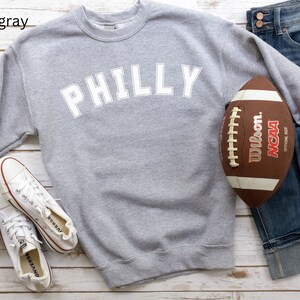 May include: A gray sweatshirt with the word "PHILLY" printed in white on the front. The sweatshirt is laid out on a white wooden surface with a football and a pair of white sneakers.