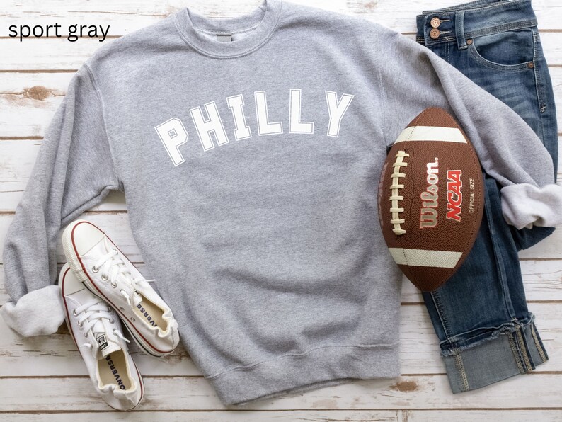 May include: A gray sweatshirt with the word "PHILLY" printed in white on the front. The sweatshirt is laid out on a white wooden surface with a football and a pair of white sneakers.