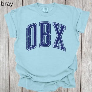 May include: A light blue t-shirt with the text "OBX" in navy blue, distressed lettering. The shirt is made of a soft material and has short sleeves. The word "Chambray" is in the top left corner.