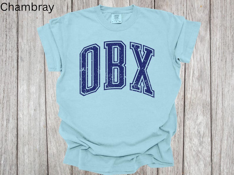 May include: A light blue t-shirt with the text "OBX" in navy blue, distressed lettering. The shirt is made of a soft material and has short sleeves. The word "Chambray" is in the top left corner.
