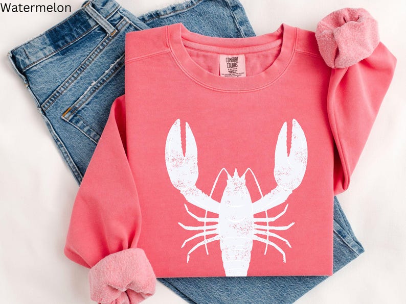 May include: A pink sweatshirt with a white lobster graphic. The sweatshirt is folded and laid on a pair of blue jeans.