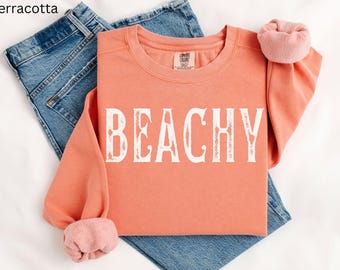 May include: A terracotta colored sweatshirt with the word "Beachy" printed in white on the front. The sweatshirt is folded and laid on top of a pair of blue jeans.