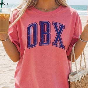 May include: A pink t-shirt with the letters "OBX" in a distressed navy blue collegiate font. The shirt is worn by someone holding a drink and a straw bag. The word "Watermelon" is in the top left corner.