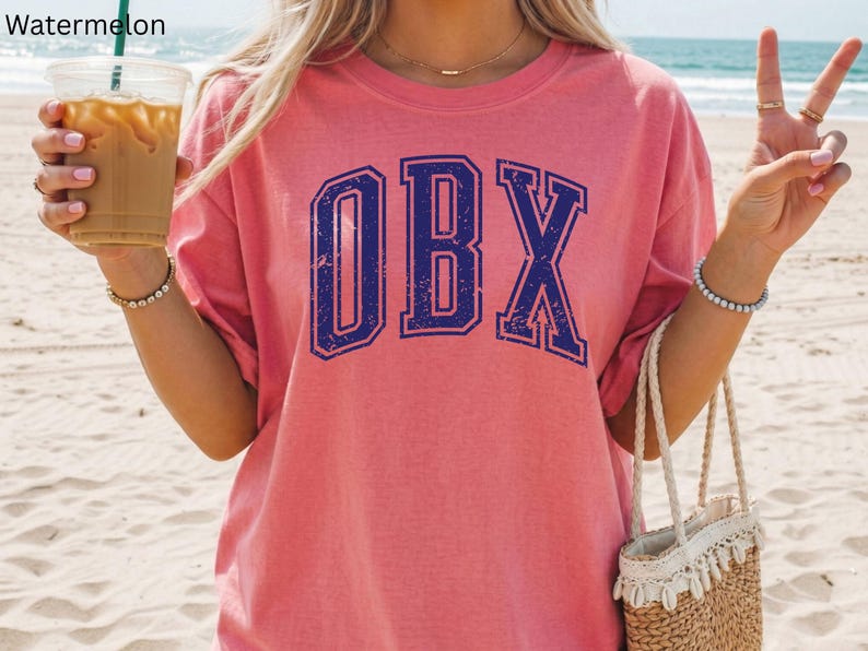 May include: A pink t-shirt with the letters "OBX" in a distressed navy blue collegiate font. The shirt is worn by someone holding a drink and a straw bag. The word "Watermelon" is in the top left corner.