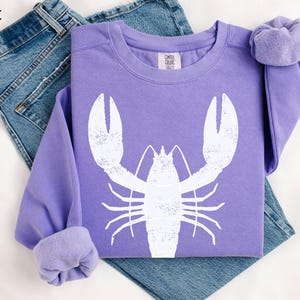 May include: A purple sweatshirt with a white lobster graphic. The sweatshirt is folded and laid on a pair of blue jeans.
