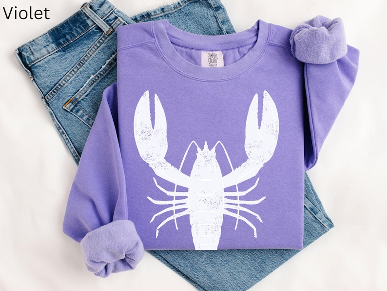 May include: A purple sweatshirt with a white lobster graphic. The sweatshirt is folded and laid on a pair of blue jeans.