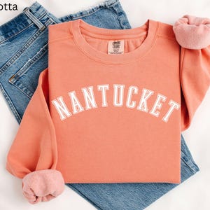 May include: A terracotta colored sweatshirt with the word "NANTUCKET" printed in white on the front. The sweatshirt is folded and laid on top of a pair of blue jeans.