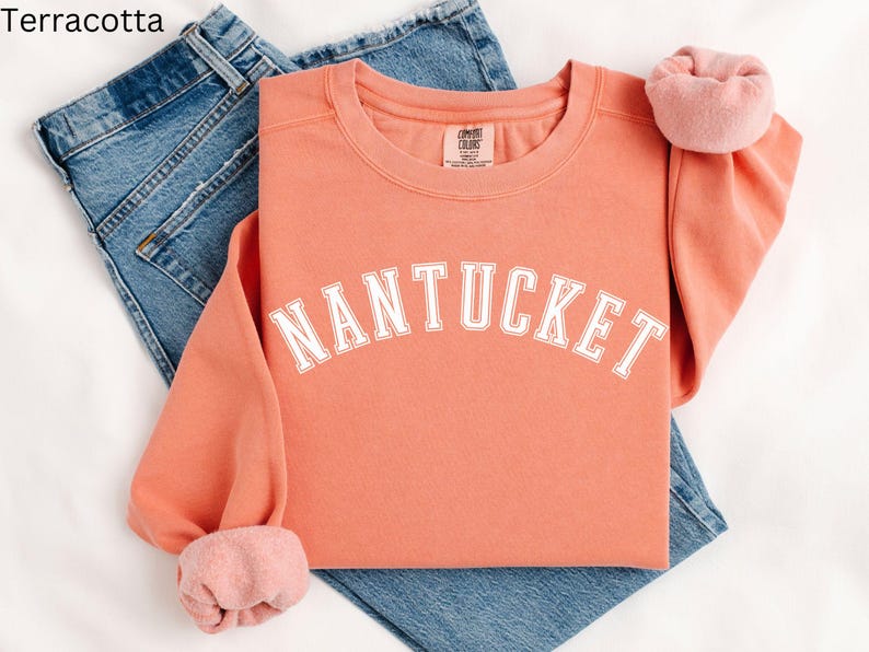 May include: A terracotta colored sweatshirt with the word "NANTUCKET" printed in white on the front. The sweatshirt is folded and laid on top of a pair of blue jeans.