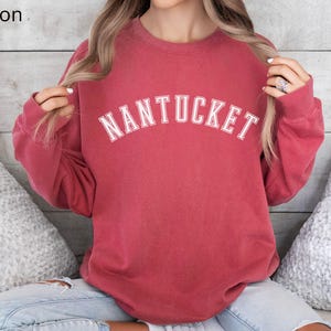 May include: A crimson red sweatshirt with long sleeves and a white crew neck. The sweatshirt has the word "NANTUCKET" printed in white on the front.