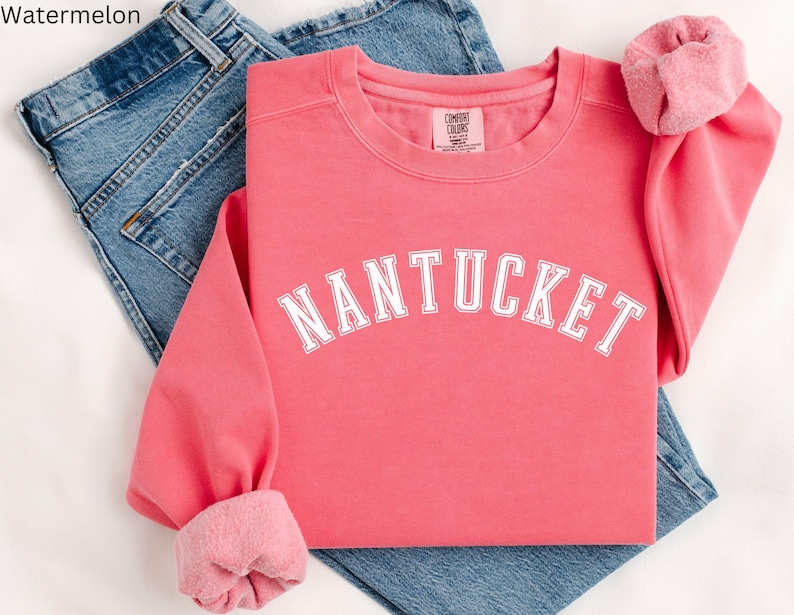 May include: A pink sweatshirt with the word "NANTUCKET" in white text on the front. The sweatshirt is folded and laid on top of a pair of blue jeans.