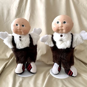 May include: Two Cabbage Patch Kids dolls wearing brown and white outfits with red and white shoes. The dolls are standing on white bases.