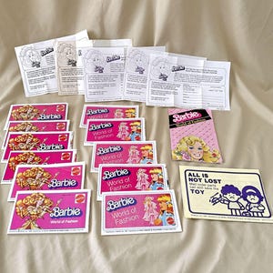 May include: A collection of pink and white cards with the text "Barbie World of Fashion" and images of Barbie dolls. There is also a pink and white booklet with the text "Barbie World of Fashion" and a white card with the text "All is not lost. Mail order parts can still save the toy."