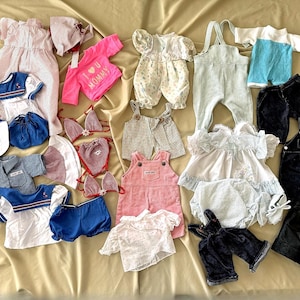 May include: A collection of doll clothes, including dresses, shirts, pants, overalls, and swimwear. The clothes are in various colors and patterns, including pink, blue, white, red, and plaid.
