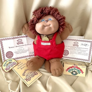 May include: A brown Cabbage Patch Kid doll with brown hair and blue eyes wearing a red overall and a gold necklace. The doll is sitting on a beige fabric surface with a certificate, a box, and two tags that say "Cabbage Patch Kids" and "Koosas" in front of it.