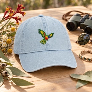 Puede incluir: Una gorra de béisbol de mezclilla azul claro con un gráfico bordado de un loro colorido con plumas verdes, amarillas y rojas en vuelo. La gorra tiene una visera curva, un botón en la parte superior y ojales cosidos para ventilación.
