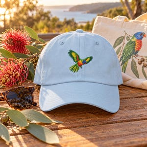 Peut inclure: Une casquette de baseball bleu clair avec une broderie représentant un perroquet coloré en plein vol sur le panneau avant. La casquette présente une visière incurvée, un bouton sur le dessus et des œillets cousus pour la ventilation, avec des plumes vertes, jaunes et rouges.